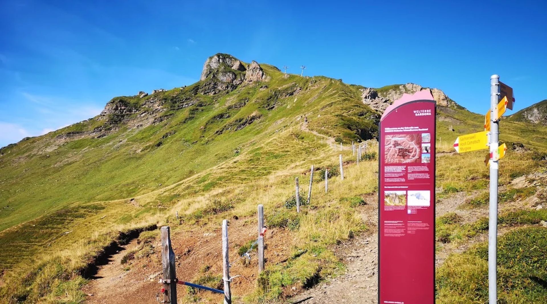 Hiking trail at Pizol, St. Gallen, with a UNESCO World Heritage Sardona information panel and a yellow Swiss hiking signpost, mountain ridge with lift masts in the background